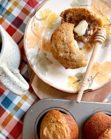 Bran muffin open with melted butter pat, honey drizzled on fall plaid tablecloth with coffee mug